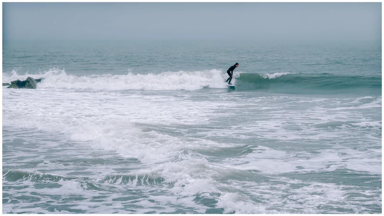 Surfer in a full wetsuit riding a small wave on a foggy day at Cape May, New Jersey