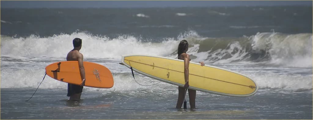 Two surfers holding longboards in the shorebreak at Folly Beach, South Carolina, watching a set roll in