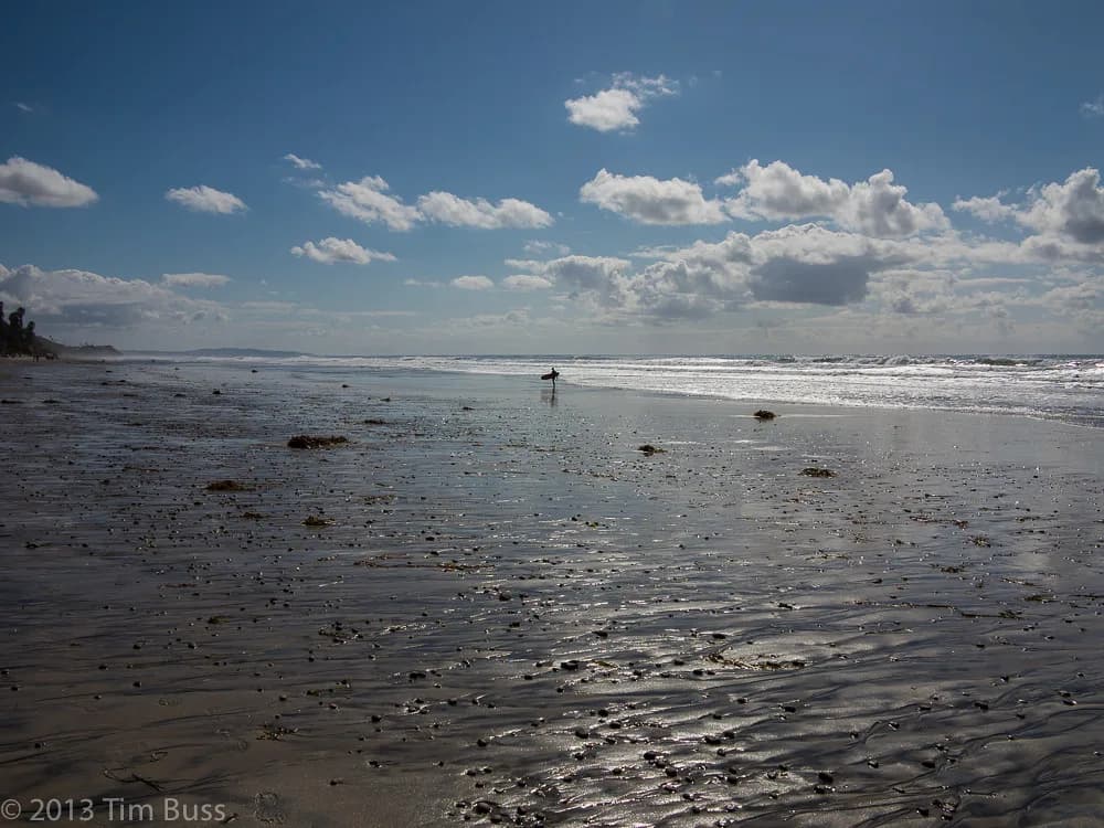 South Ponto Beach in Carlsbad at a minus tide, with a wide wet sand flat and one surfer walking to the water